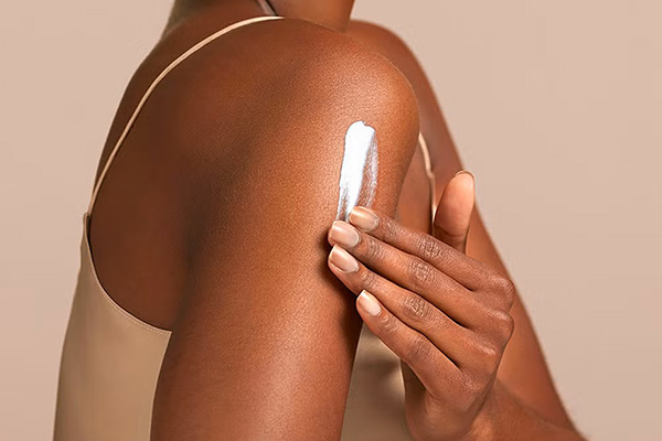 a dark skinned model applying a cream onto her shoulders. shot in a studio, you cannot see her face just her shoulders and hands