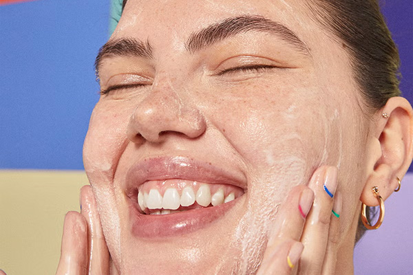 A close up image of a female model rubbing her face with BYOMA’s Jelly cleanser, in front of a multicoloured background.