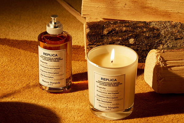 A image of Maison Margiela Replica By The Fireplace fragrance and candle sat in front of fire wood on a orange background.