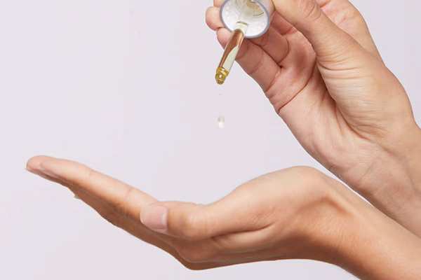 A medium shot of a female hand putting droplets of serum in her hand, in a studio setting, in front of a white background.