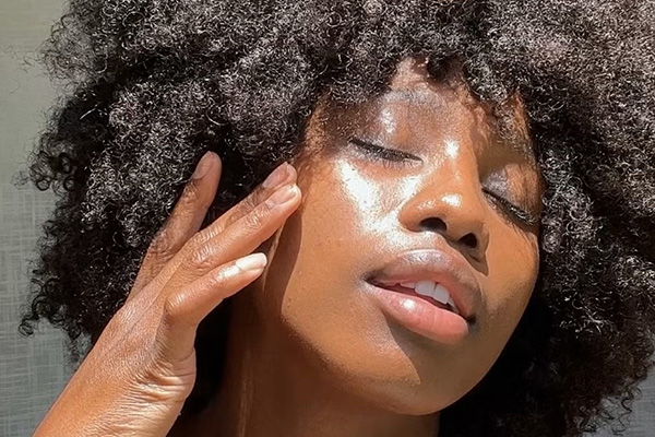 Dark skin model closing her eyes and touching her cheeks. the sun on her face exposing her glowy skin. model has afro hair