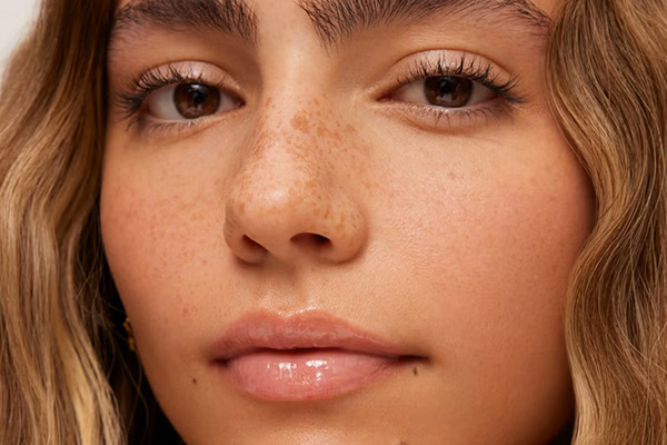 model staring into the camera. she has freckles, dark eyes and glossy lips. her hair is golden and her eyes are brown.