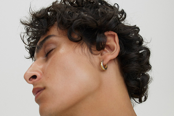 A close up of a male model with dark brown curly hair looking away from the camera, in a studio setting, in front of a white background.