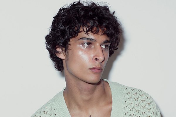 A close up of a male model with dark brown curly hair looking away from the camera, in a studio setting, in front of a white background.