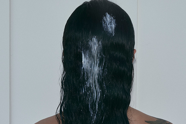 A medium shot of the back of a female’s wavy, brunette hair as she is in the shower applying her smoothing shampoo from Hello Klean.