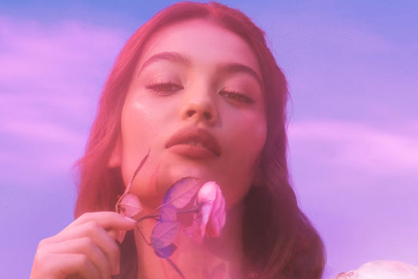 a pink and purple tinted image of a model with brown hair holding a pink rose next to her chin. she stands against a lavender sky 