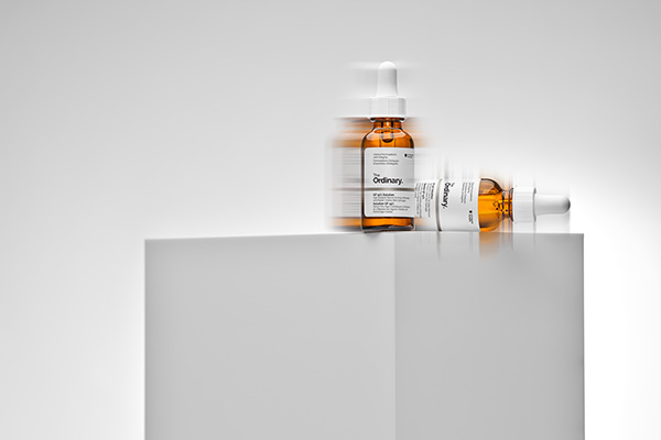 A blurred image of The Ordinary’s Growth Factors Solution 15% Serum on a tall white cylinder, in a studio setting in front of a white background.