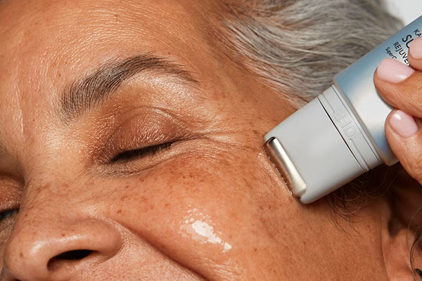 a medium skinned older model using a serum on her cheeks. her eyes are closed, her hair is grey. the image is a close up of her cheeks and eyes