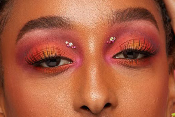 a medium skinned model with an aura make up look on her eyes using oranges and purples. she has glitter pieces on each eye