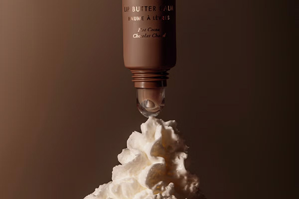 A close up of Summer Friday’s Hot Chocolate lip butter balm sitting on top of a pile of whipped cream, in front of a brown background.