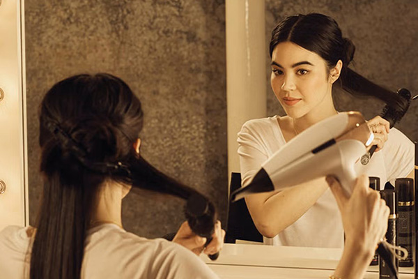 a brown haired model sitting infront of a mirror blow drying her hair using a white ghd hair dryer