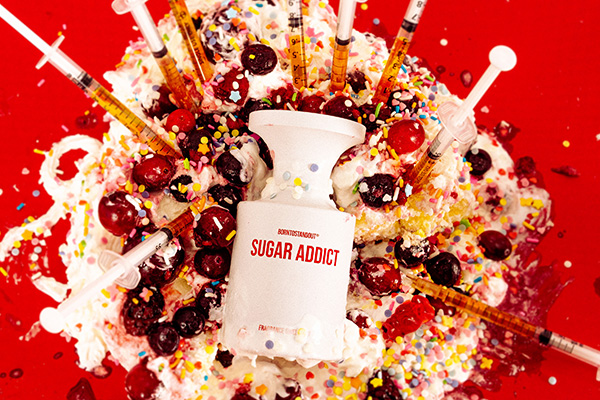 A image of BORNTOSTANDOUT's Sugar Addict perfume bottle splashed on a cherry cake with a medical injections as candles.