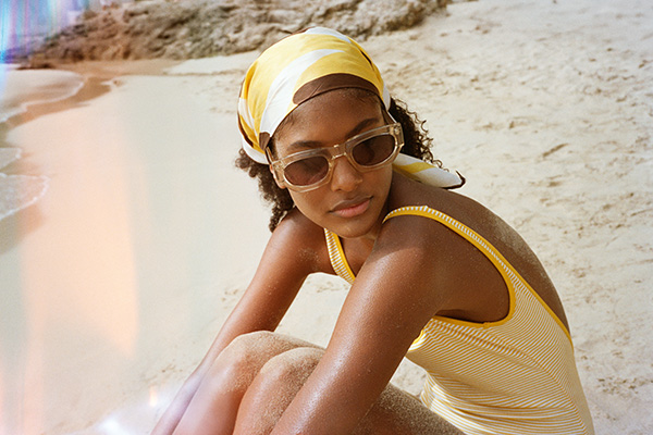 a image of a female model wearing a yellow swimsuit, sat on the sand of a beach with sunglasses looking away from the camera.
