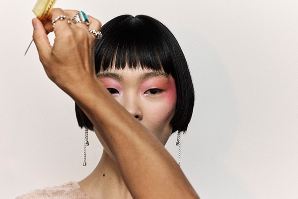A model with a black bob and fringe having her hair fixed, the stylists hands cover halt her face and hse has bright pink eyeshadow on