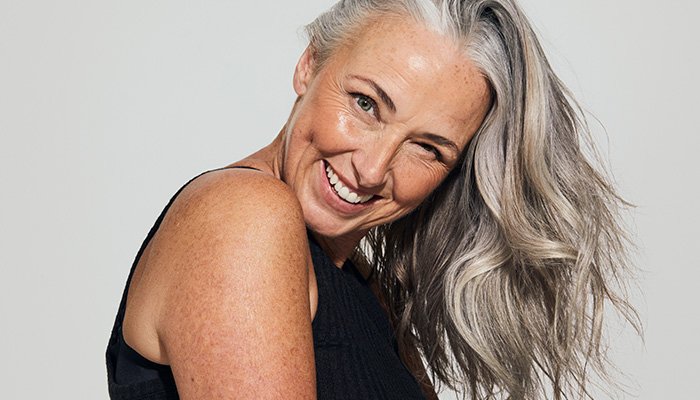 A medium shot of older model with grey hair smiling at the camera in a studio setting on a grey background.