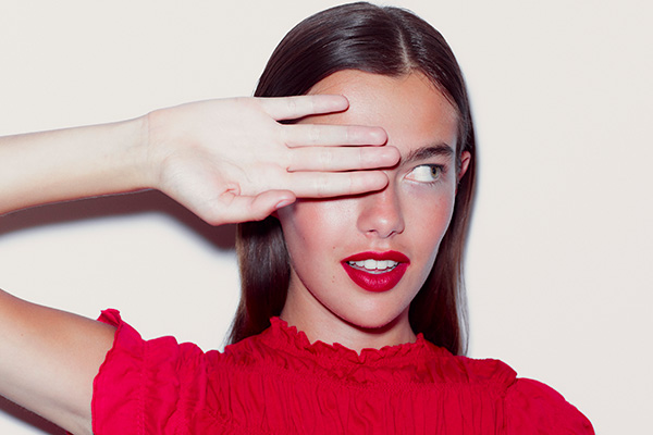 A medium shot of a woman wearing a red top and red lipstick covering one of her eye with her hand – in a studio setting in front of a white background.