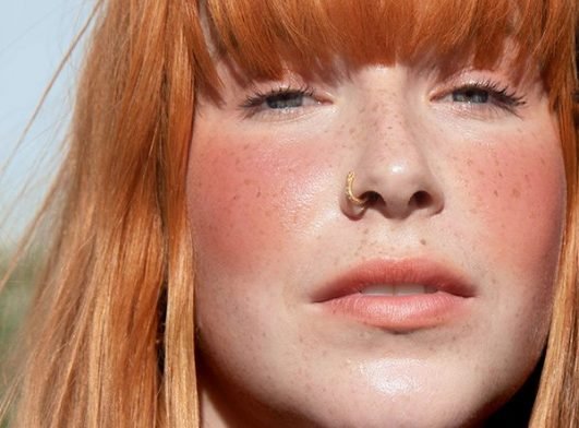 Red headed model with fair skin, blue eyes and a gold nose ring gazes at the camera, wearing a peach-hued blusher on her cheeks.