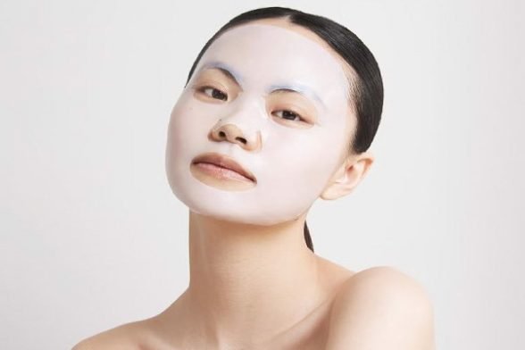 A meduim shot of a Asian female model wearing a Decree sheet face mask and looking into the camera on a white background.
