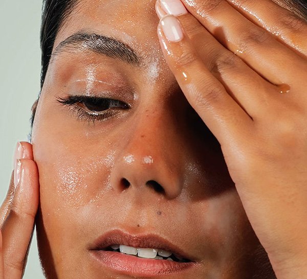A close up image of a woman rubbing in a barrier-boosting serum into her face as she looks down in a studio setting, on a grey background.