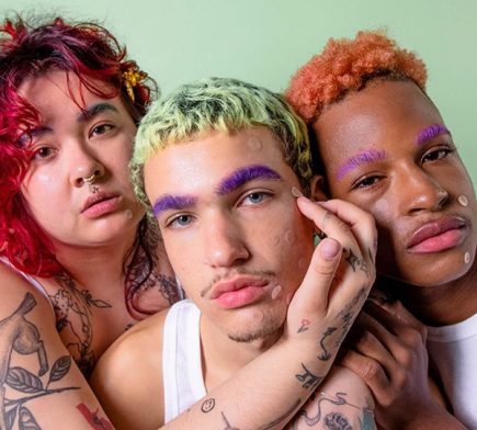 A medium shot of three young models with coloured hair and eyebrows wearing see-through pimple patches on a green background.