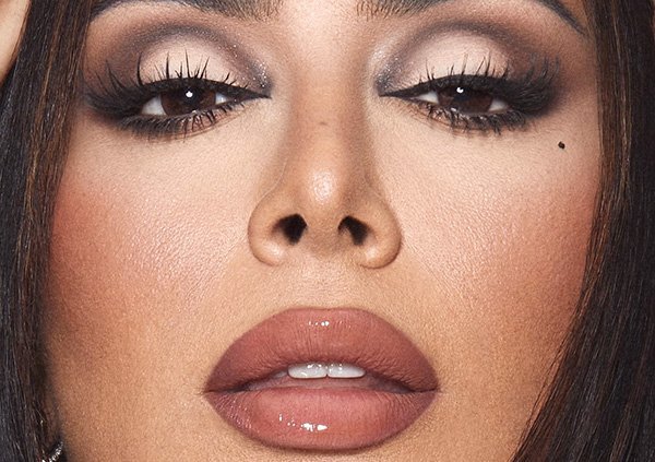A close up image of a woman with dark hair wearing full coverage foundation, smoky eye make up, false lashes and a nude glossy lip.
