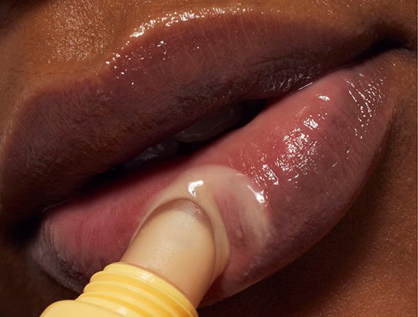 A very close up image of a dark-skinned person applying a moisturising lip balm from a yellow applicator directly onto their lips