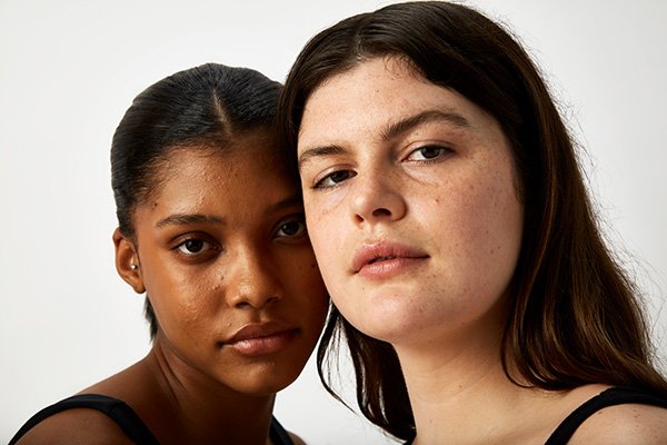 Two female models in a studio setting against a white background look at the camera, they both have dark hair and wear no make up