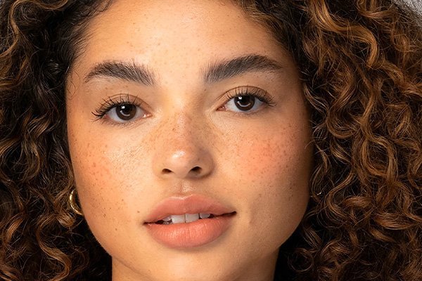 A close up of a model with brown curly hair, brown eyes, groomed brows, freckles and peachy lips looking straight at the camera.