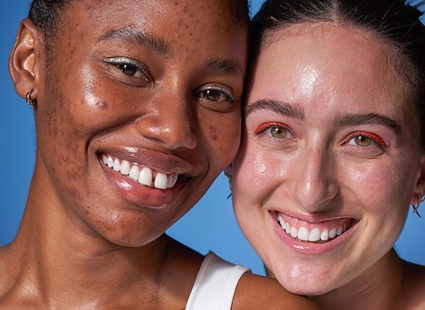 Two female models with bare skin and minimal eye make up pose side by side in a studio setting against a light blue background