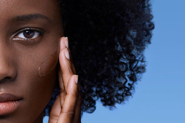 a dark skinned model with an afro touching her cheek, shot against a blue background. the image is just half of the models face.