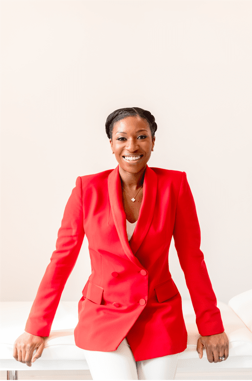 A black skincare specialist is perched on a white clinic bed smiling at the camera and wearing a bright red blazer 