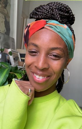 A selfie of smiling black woman wearing a green jumper, a red and green headscarf and braided hair in a high bun