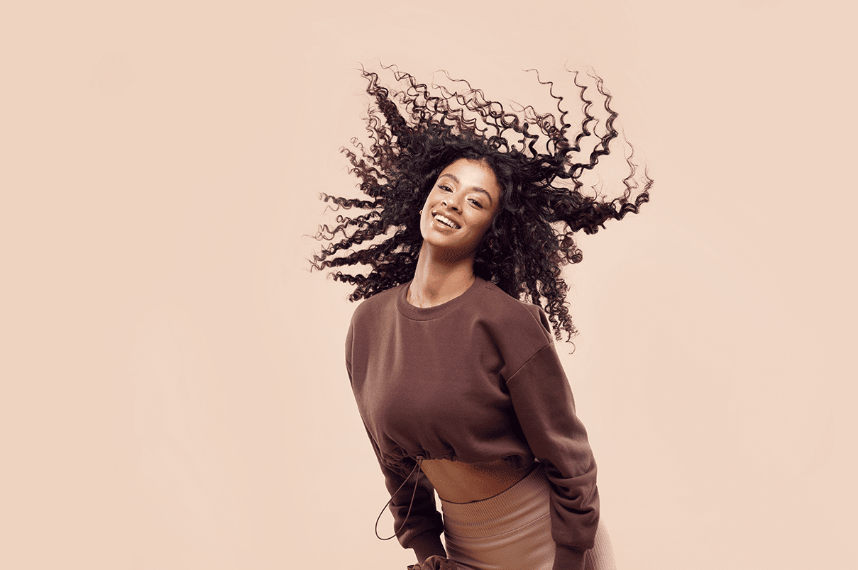 Curly haired woman, positively smiling while doing a hair flick, brown background.