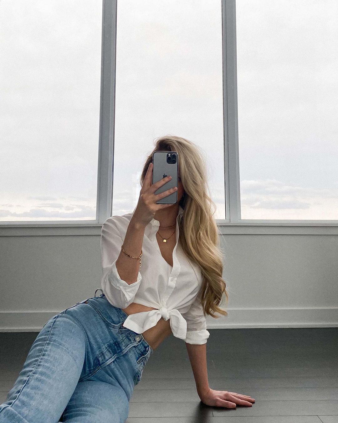 Woman sitting down wearing jeans and white shirt with long blonde hair, taking a picture in a mirror.