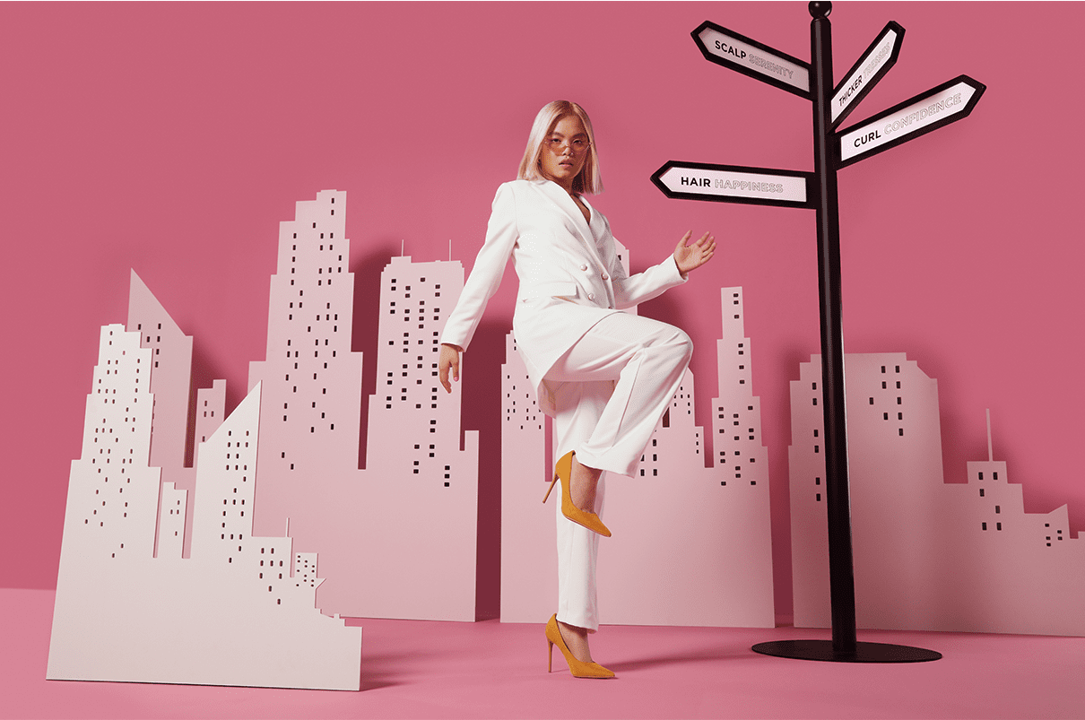 Woman with blonde straight hair, wearing a pink suit walking past a pink city skyline.