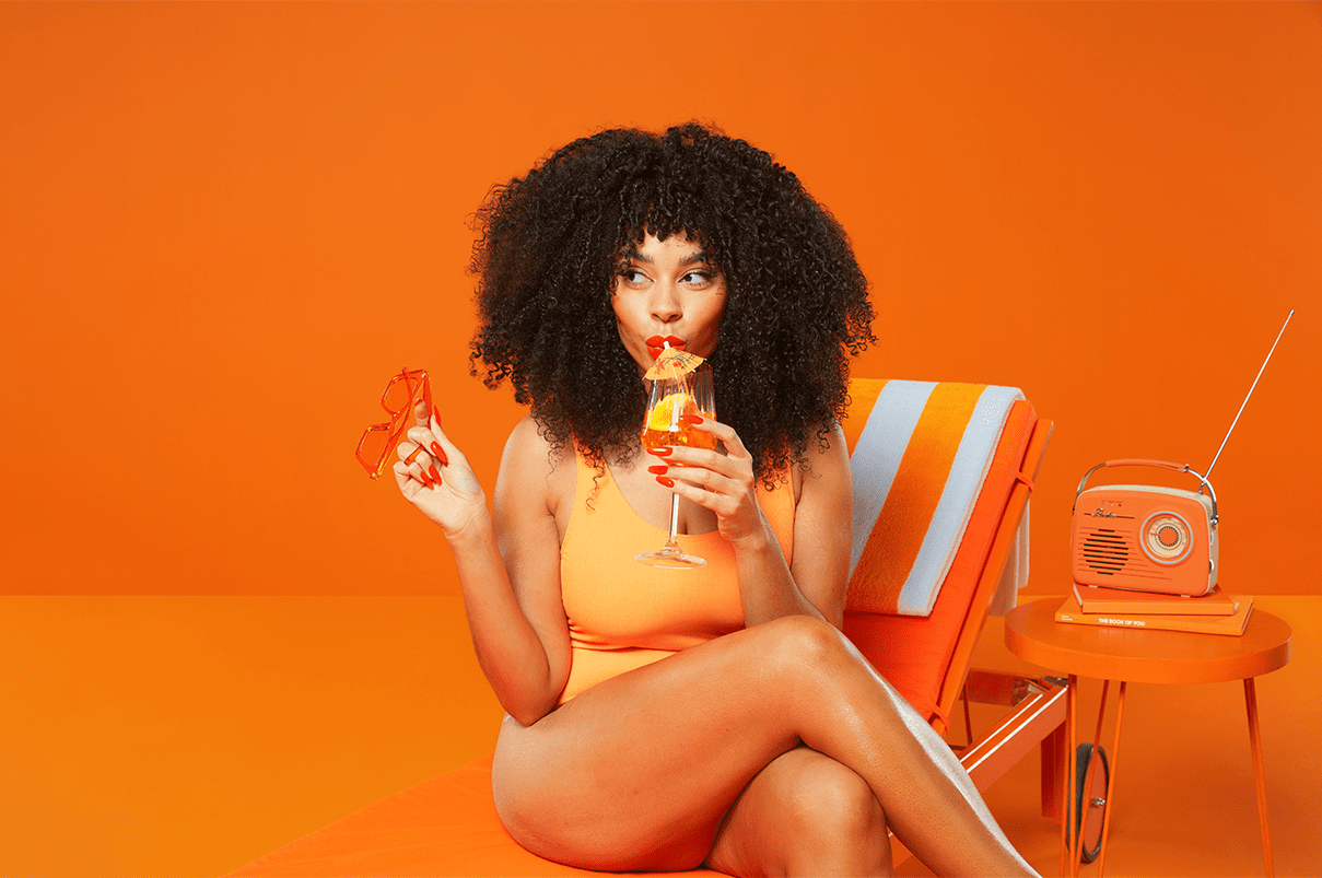 Woman with curly hair in orange swimsuit sitting on a lounger drinking a cocktail.