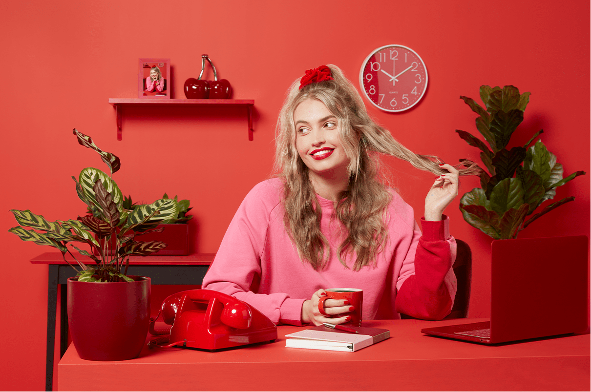 Woman with blonde wavy hair sitting in her red home office surrounded by plants and a laptop.
