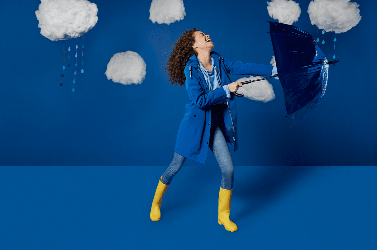 Woman with long curly hair wearing a blue raincoat and yellow welly's, holding up a blue umbrella fighting the rain.