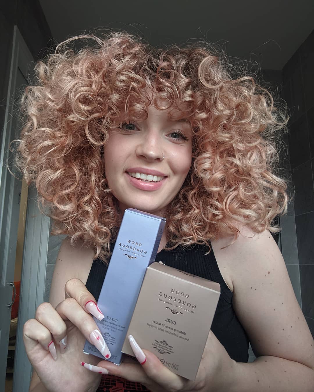 Woman with short blonde curly hair holding up two Grow Gorgeous products; the curl butter and repair oil.