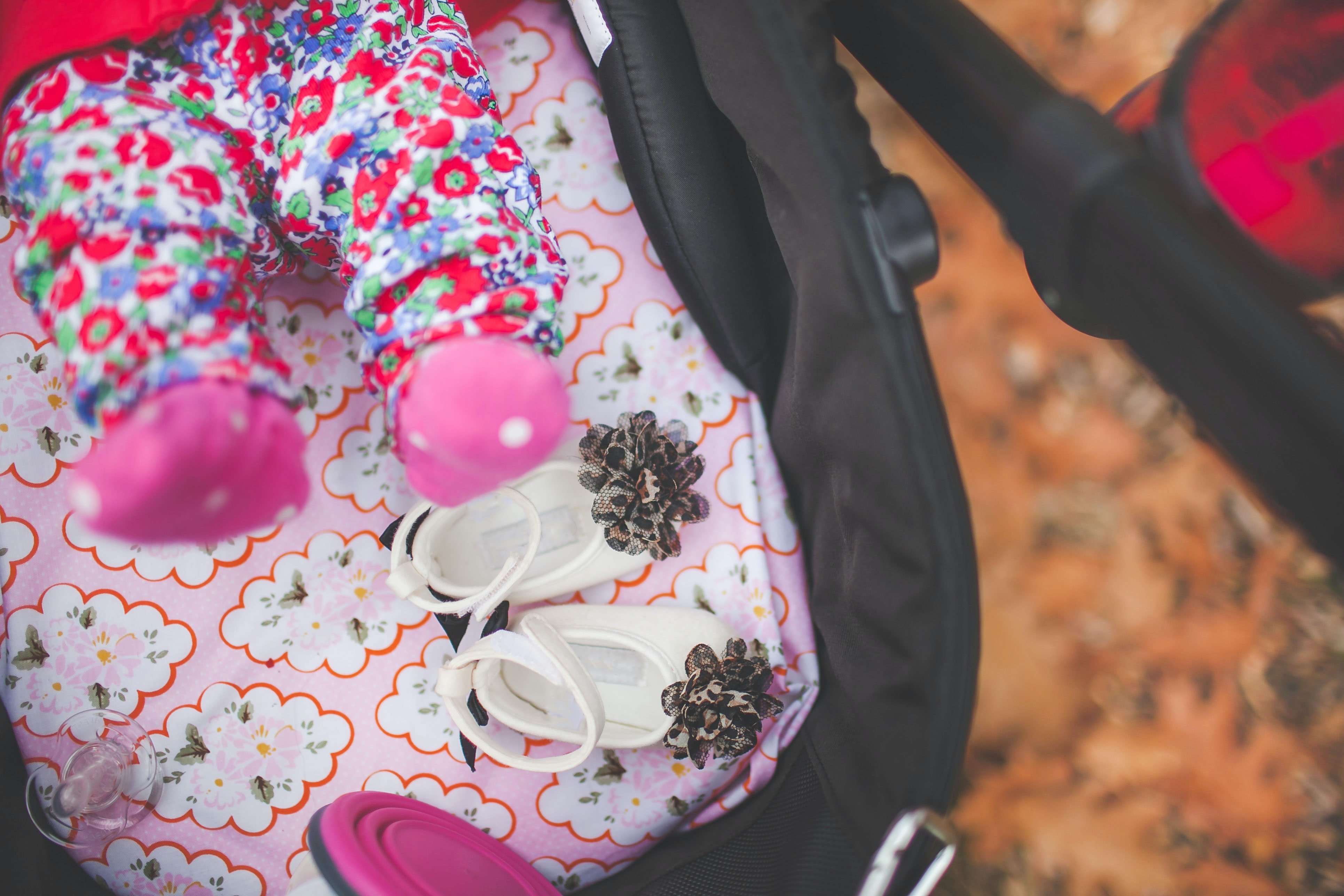 baby lying on a pink blanket in a pram outside. There are orange leaves on the floor below. Baby is wearing colourful floral bottoms and pink socks. Pram also has two baby shoes, a pacifier in it.