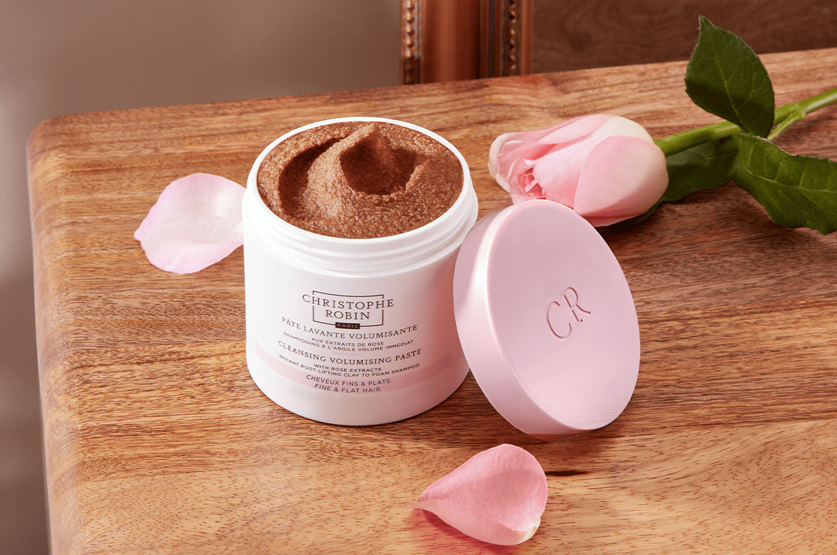 White container of the Cleansing Volumising Paste with Rose Extracts on top of a wooden table with a pink rose next to it.