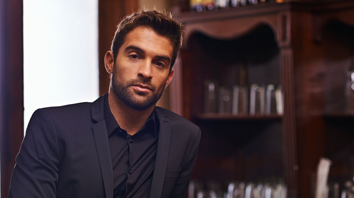 Suave, suited man with a Hollywoodian beard reclines against a dark wooden bar