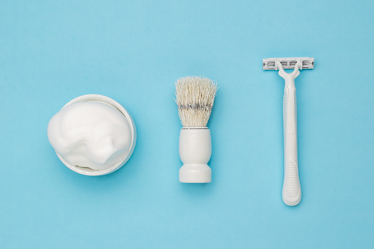 A white pot of shaving foam, shaving brush and a razor against a pale blue background | Venus UK