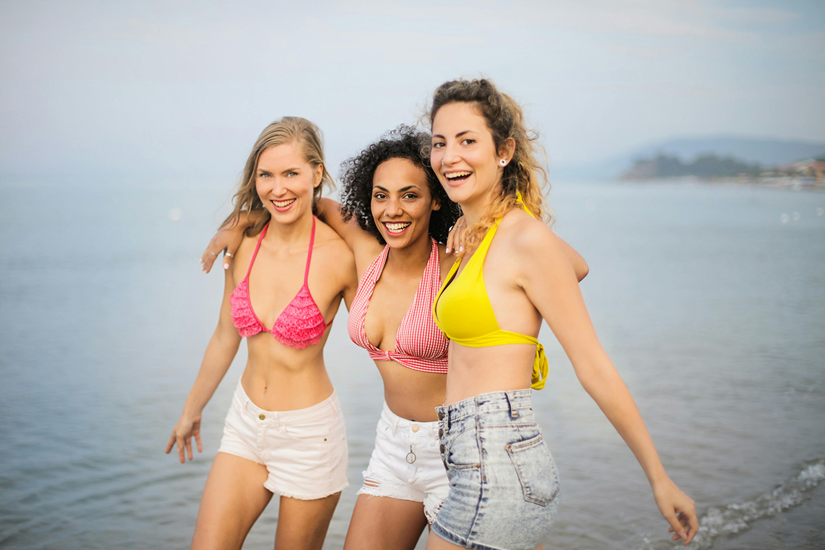 Three women in shorts and bikini tops smile on a beach | Venus UK