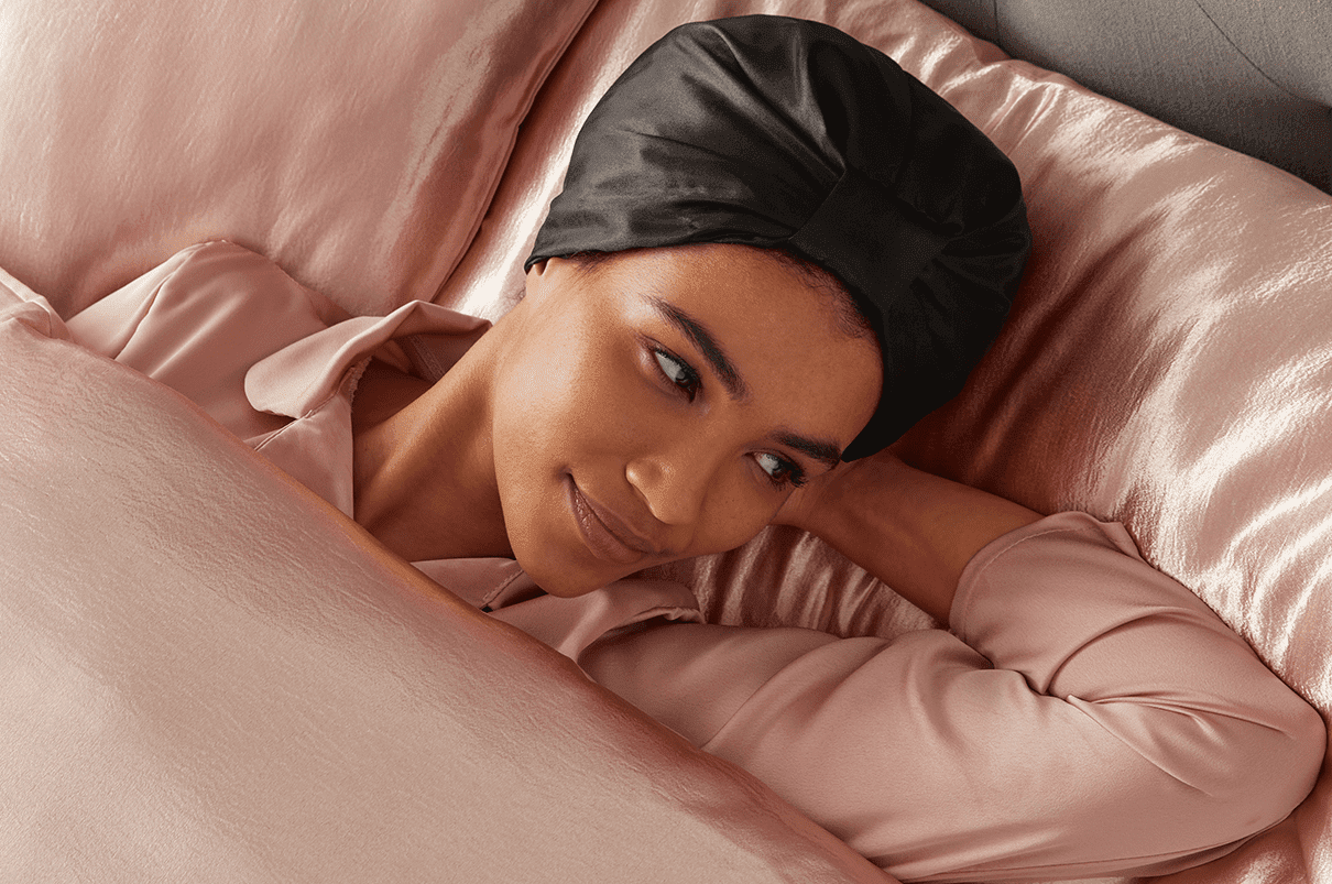 Woman laying in bed with pink bedding wearing a satin head wrap for sleeping.