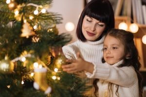 A woman wearing a Christmas eyeshadow look hangs decorations on a tree with her daughter | No7 Beauty US 