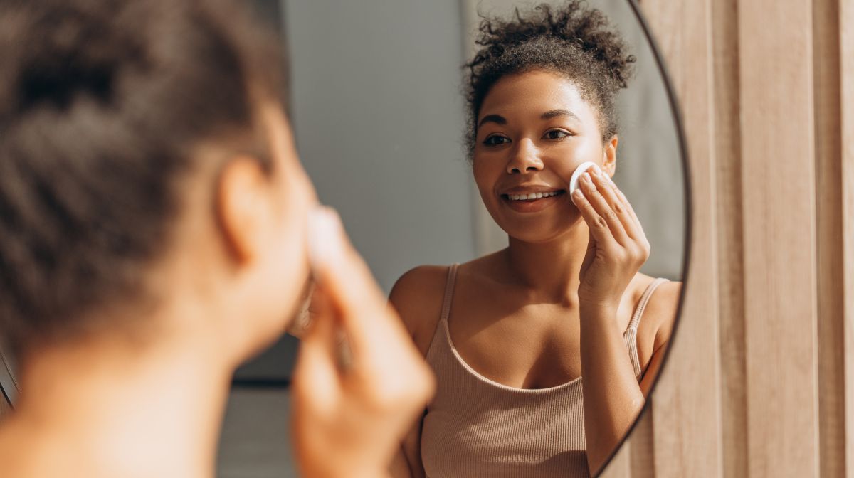 A woman uses a facial cleanser to remove her makeup in a mirror.