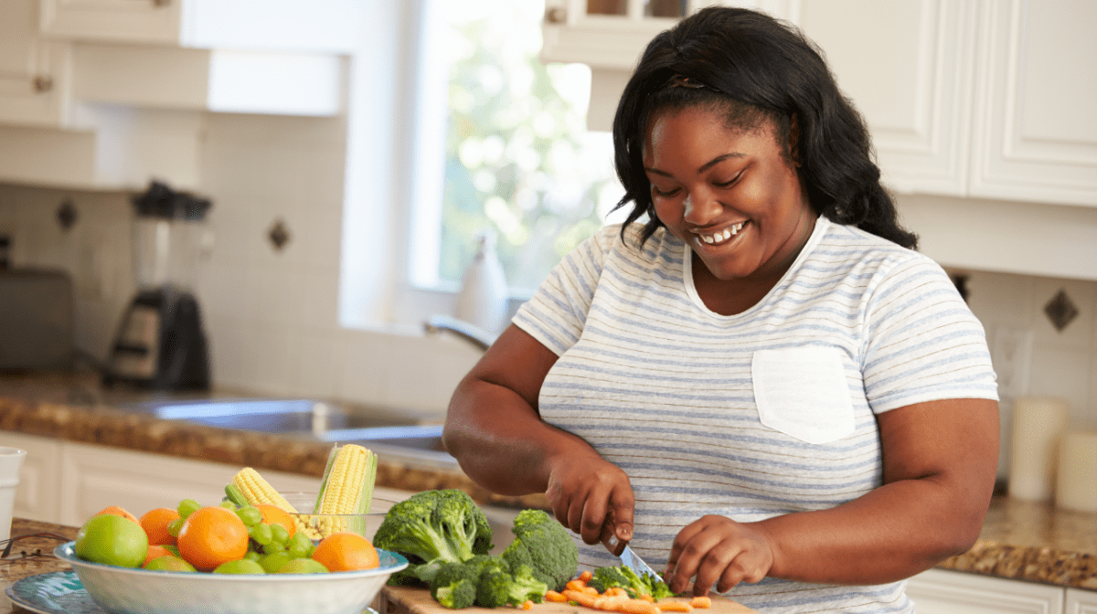 An overweight woman cutting up vegetables in a kitchen. As complex carbohydrates, vegetables fit into one of the main food groups.