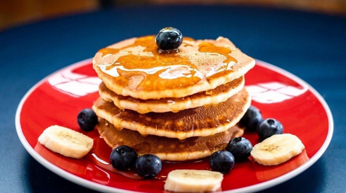 Pancakes con proteine in polvere, banane e frutti di bosco su un piatto rosso.