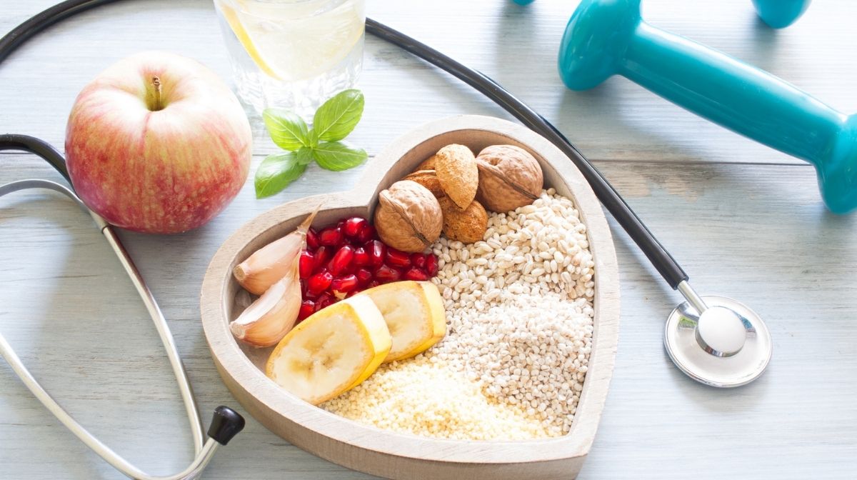 Birdseye view of heart shaped bowl containing heart healthy foods, next too dumbbells and a stethoscope.